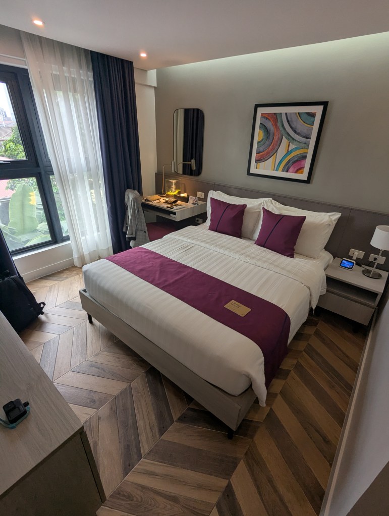 A well-decorated hotel room featuring a double bed with purple accents, a nightstand, and a small desk area. Large windows allow natural light, showcasing modern furnishings and wooden flooring.