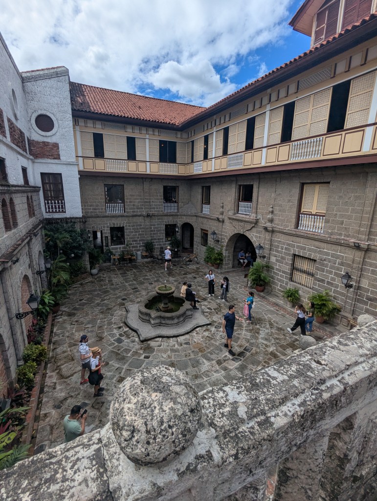 A courtyard in Casa Manila featuring a stone fountain, surrounded by lush plants and several visitors exploring the area under a partly cloudy sky.