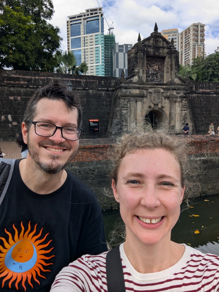 A selfie of two travelers standing in front of a historic stone structure with a decorative archway, surrounded by greenery and modern buildings in the background.