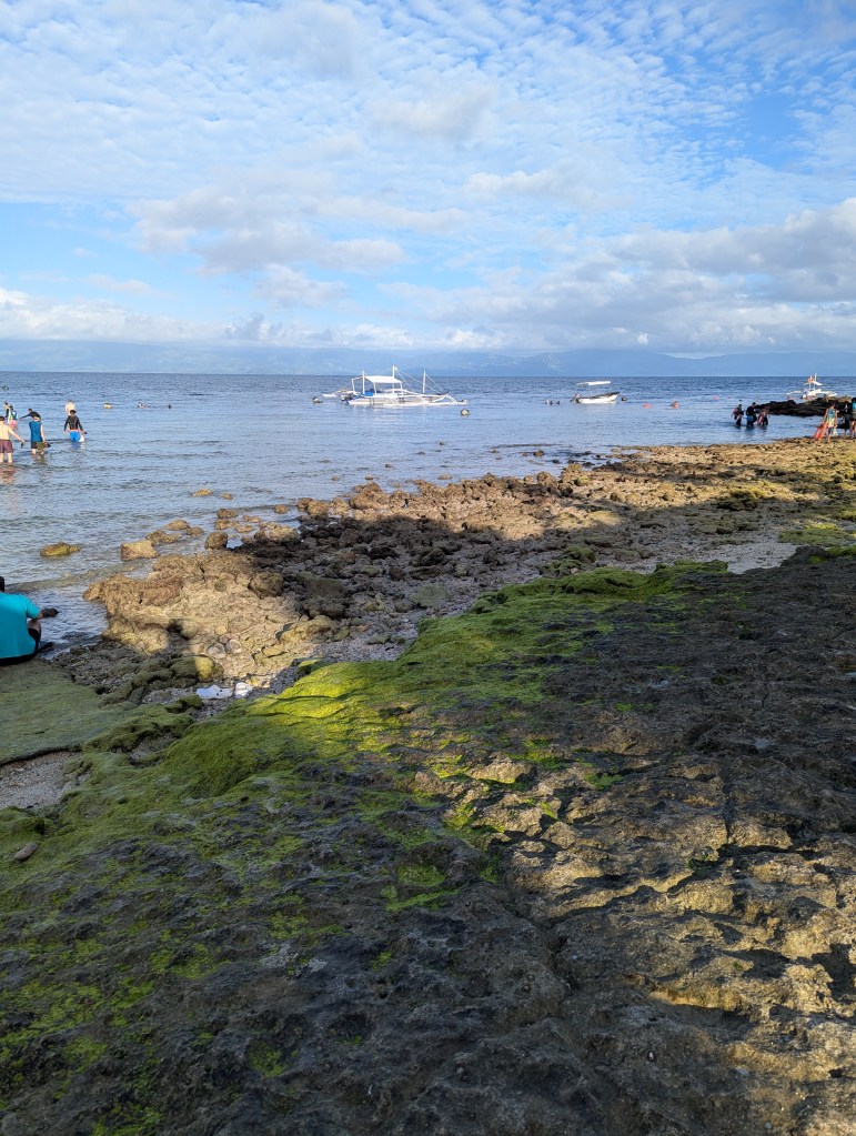 A rocky beach scene with people wading in the shallow water, a small boat anchored in the background, and a blue sky with clouds.