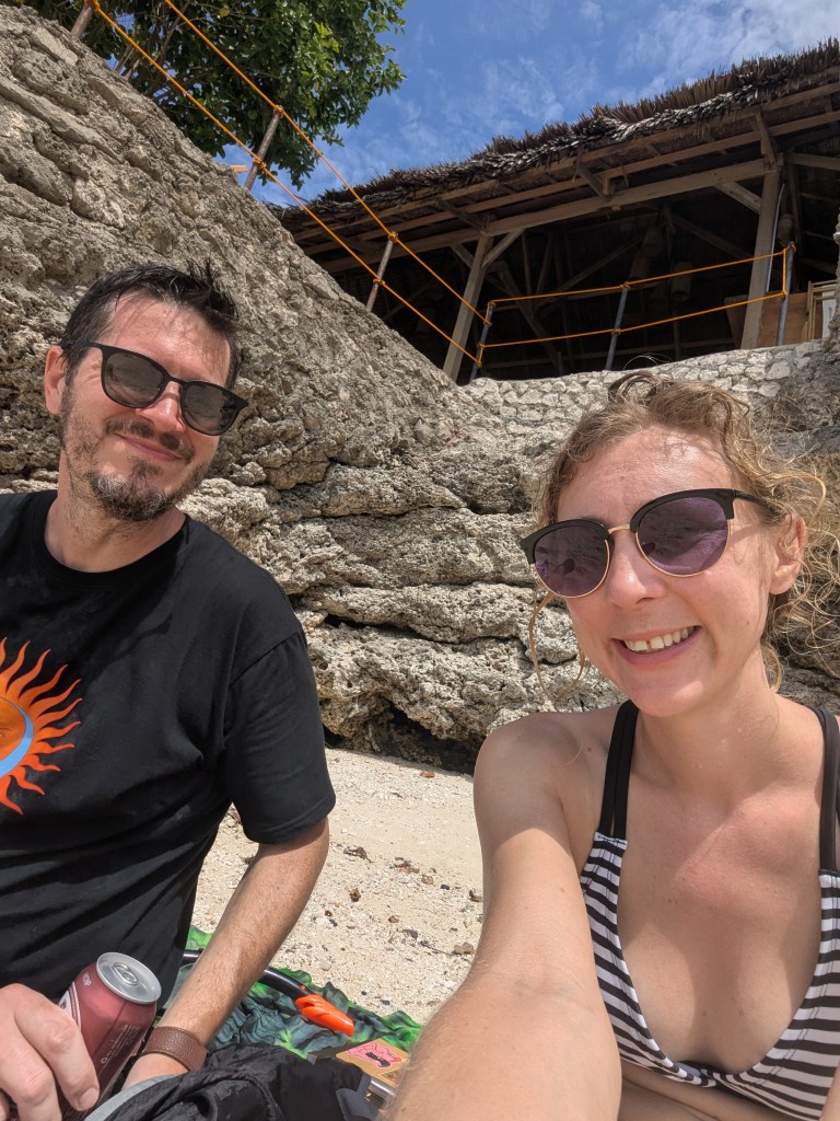 A couple sitting on a beach, enjoying drinks, with a rocky shoreline and blue sky in the background.