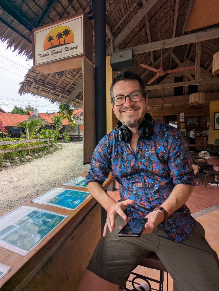 A smiling man sitting at a wooden table in front of a sign for Tipolo Beach Resort, wearing headphones and holding a smartphone.