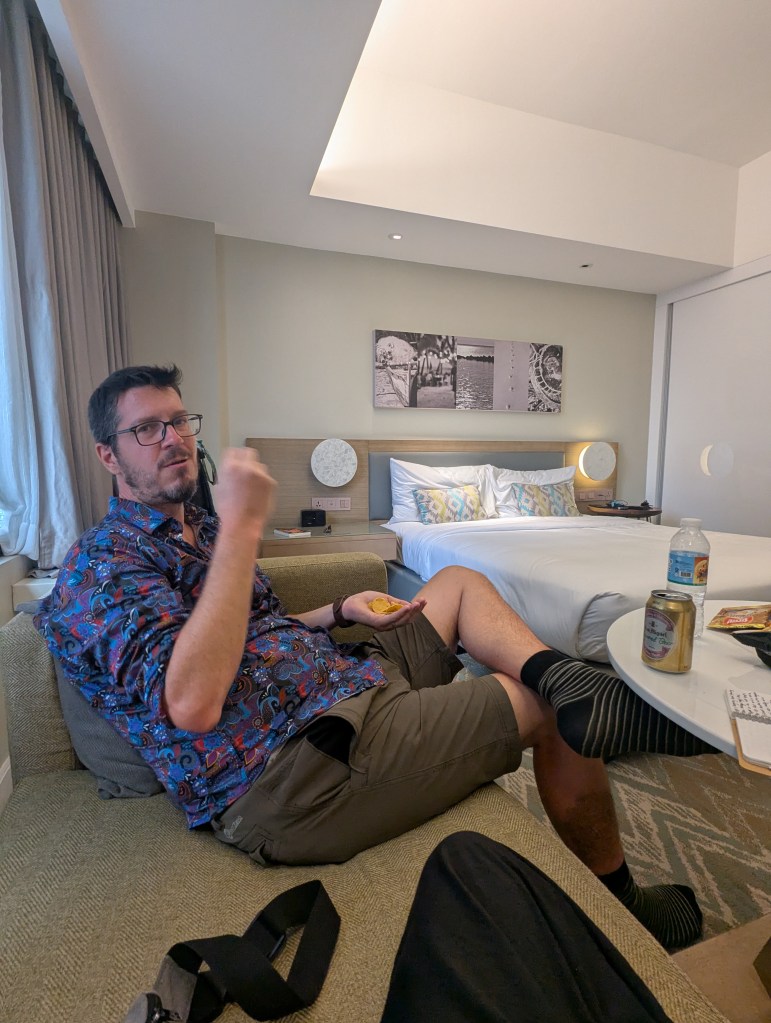 A man sitting on a couch in a hotel room, wearing a colorful patterned shirt and shorts, holding a small piece of food, with a bed and other furniture visible in the background.