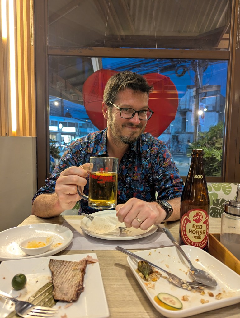 A man in a colorful patterned shirt sits at a table in a restaurant, holding a glass of beer and smiling. The table is set with several empty plates and food remnants, including fish and lime.