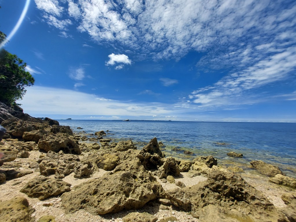 Scenic view of a rocky shoreline with clear blue ocean and sky, featuring scattered clouds.