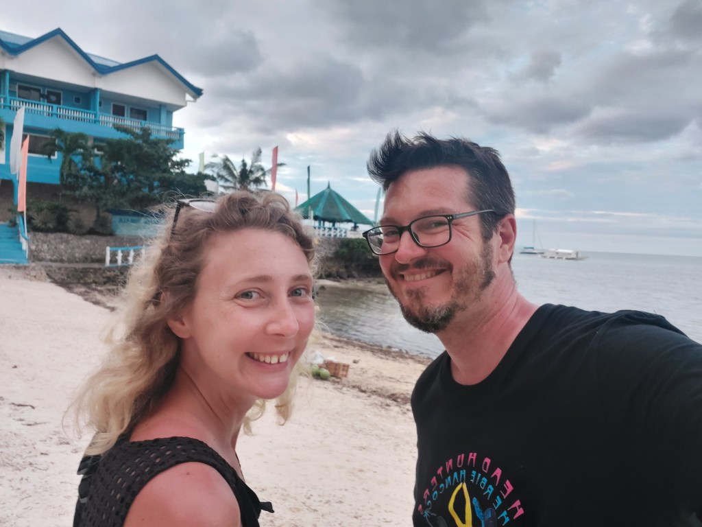 A smiling woman and man take a selfie at the beach, with a blue building and palm trees in the background under a cloudy sky.