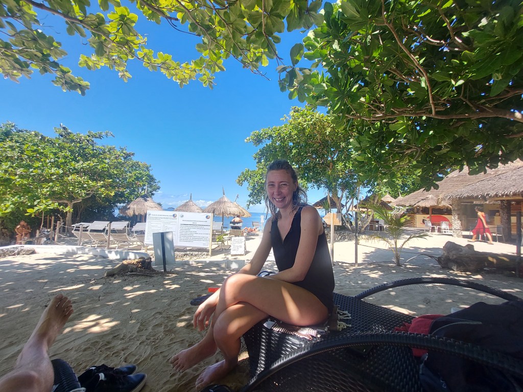 A woman sitting on a black wicker chair at a beach resort, smiling. The scene features palm trees, sun loungers, and thatched-roof huts in the background under a clear blue sky.