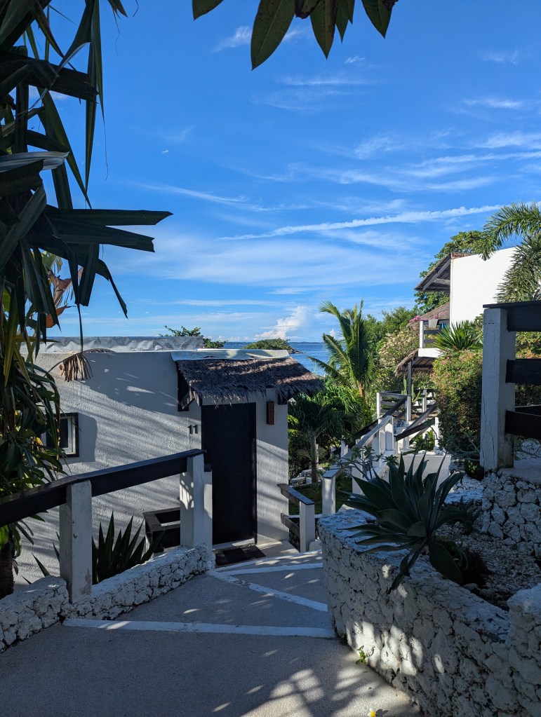 A scenic pathway leading to a small white building with a thatched roof, surrounded by lush greenery and palm trees under a blue sky.