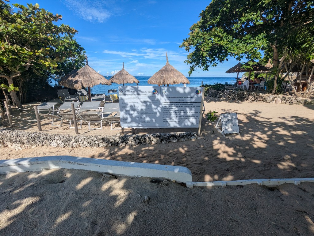 A serene beach scene featuring lounge chairs under thatched umbrellas, with a clear view of the ocean in the background. A sign with information is displayed nearby, surrounded by tropical greenery.