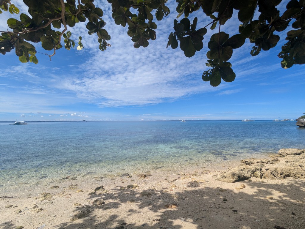 A serene beach view with clear blue water, sandy shore, and scattered rocks, framed by green leaves overhead under a bright sky with fluffy clouds.
