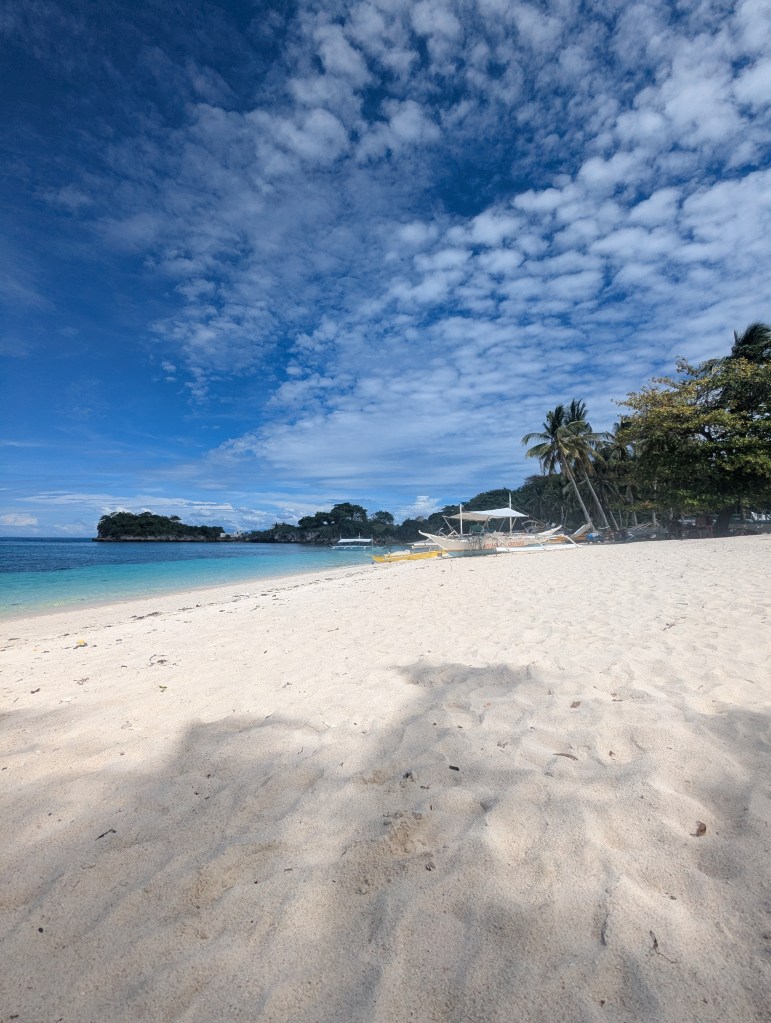 A sandy beach with clear turquoise water and a partly cloudy blue sky. Palm trees are visible on the right, and a small boat is anchored in the distance.