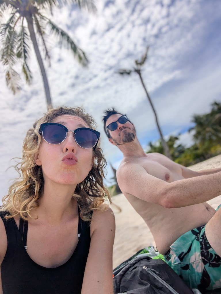 A couple enjoying a sunny day at the beach, posing for a selfie. The woman, with curly hair, is making a playful kissy face, while the man, wearing sunglasses, looks relaxed. Palm trees and a cloudy sky are visible in the background.