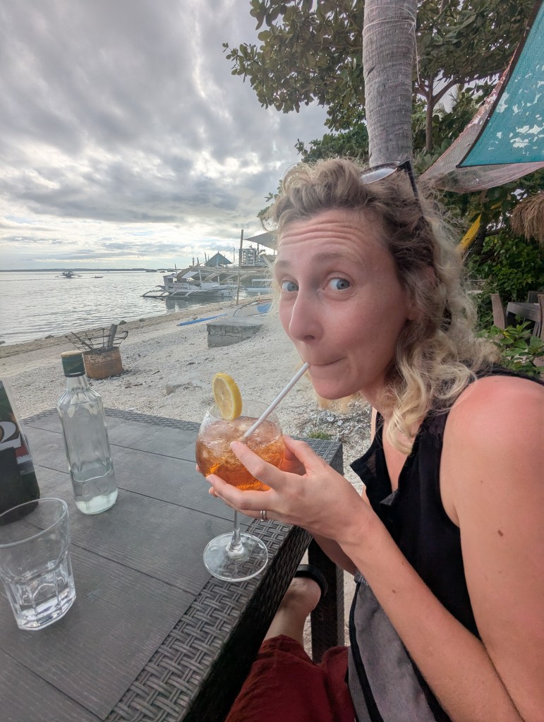A cheerful woman enjoying a drink with a slice of lemon at a beachside table, with a scenic view of the ocean and cloudy sky in the background.