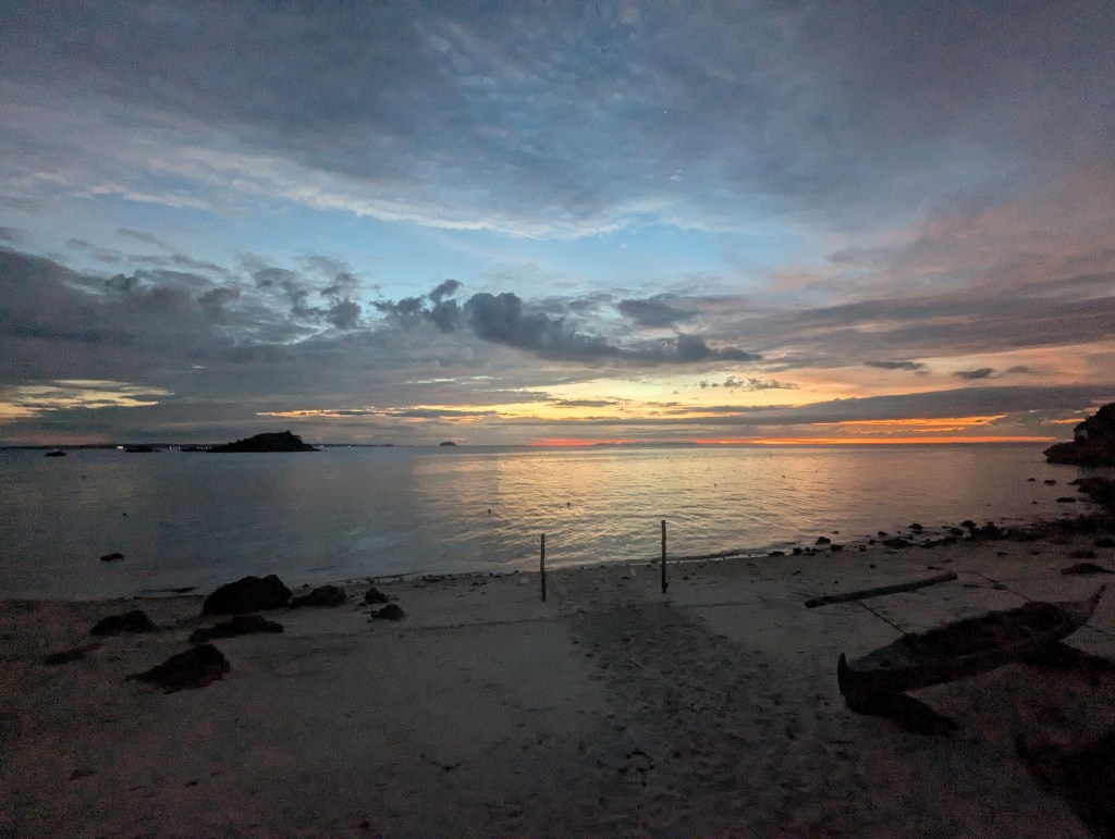 Serene beach sunset view with calm water reflecting colorful clouds and distant islands.