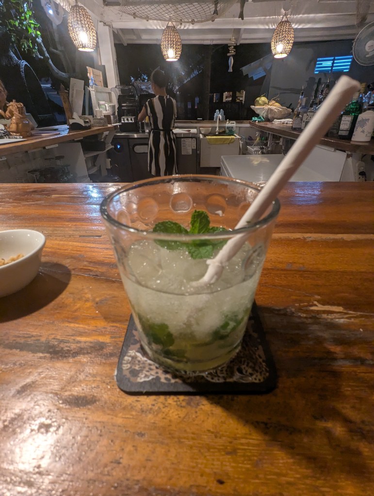 A close-up of a mojito cocktail with mint leaves and ice on a wooden bar, with a blurred figure of a bartender in the background.