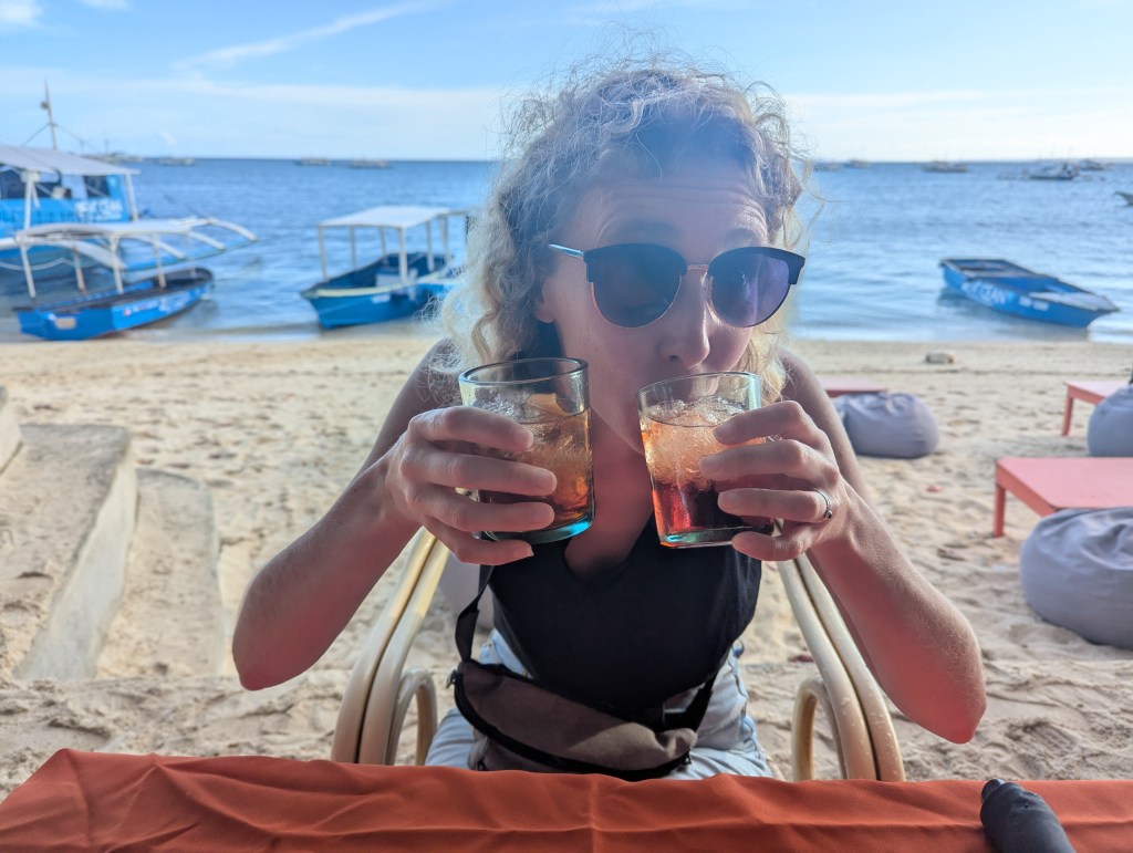 A person sitting at a beachside table holding two glasses of a dark beverage, with a view of the ocean and boats in the background.