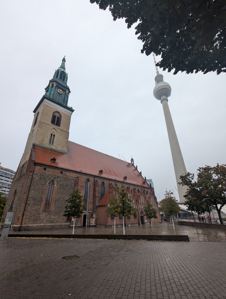 View of a historic church with a tall clock tower and the Berlin TV Tower in the background on a cloudy day.