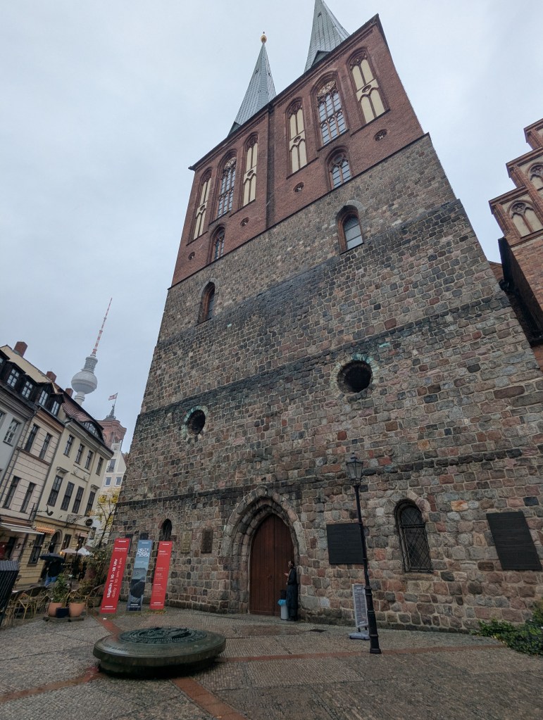 View of a historic stone building with tall spires, located in a public square, featuring a wooden door and adjacent lampposts.