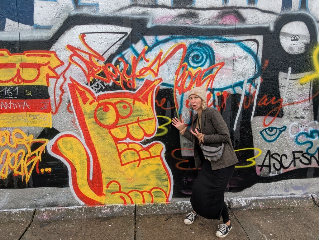A woman in a gray blazer and beanie poses playfully beside a colorful graffiti mural, featuring a large cartoon cat in yellow and red.