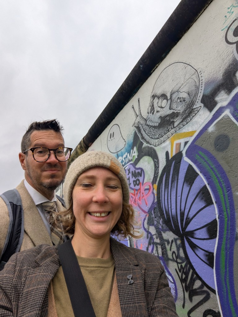 A man and a woman posing for a selfie in front of colorful street art, including a skull and graffiti, on a cloudy day.