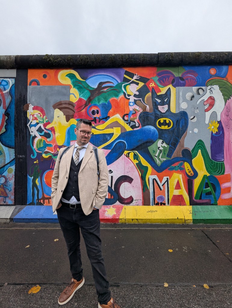 A man wearing a light blazer, vest, and glasses stands in front of a vibrant mural featuring various comic book characters and colorful abstract designs on the Berlin Wall.
