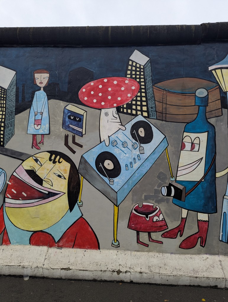 A colorful mural depicting various cartoonish characters, including a DJ, a woman in a blue dress and red boots, a large smiling man, and a mushroom, against a city backdrop.