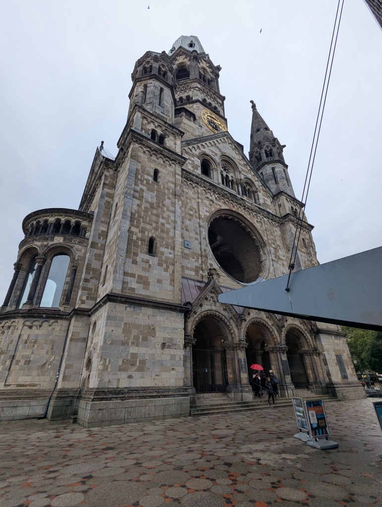 View of a historic stone church with a tall tower, featuring intricate architecture and a large round window, under a cloudy sky.