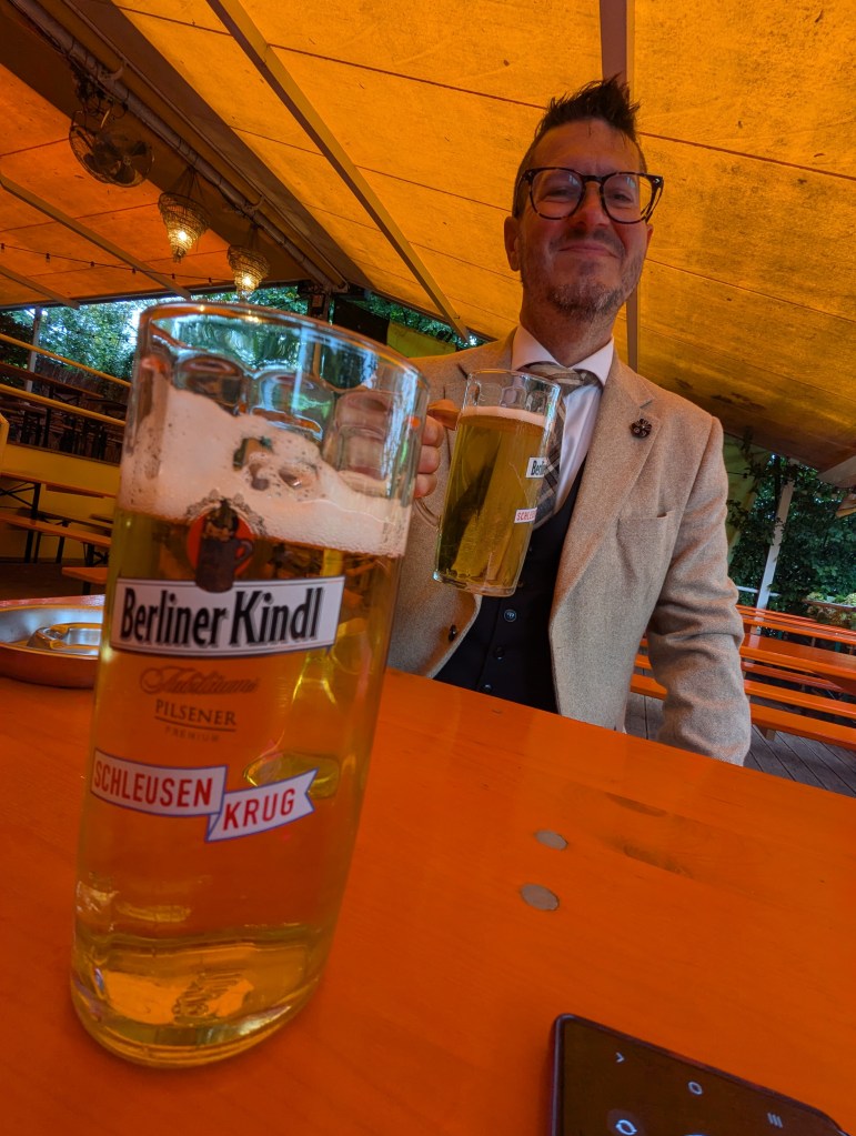 A man in a beige suit and glasses smiling while holding a beer in a lively outdoor setting with an orange canopy.