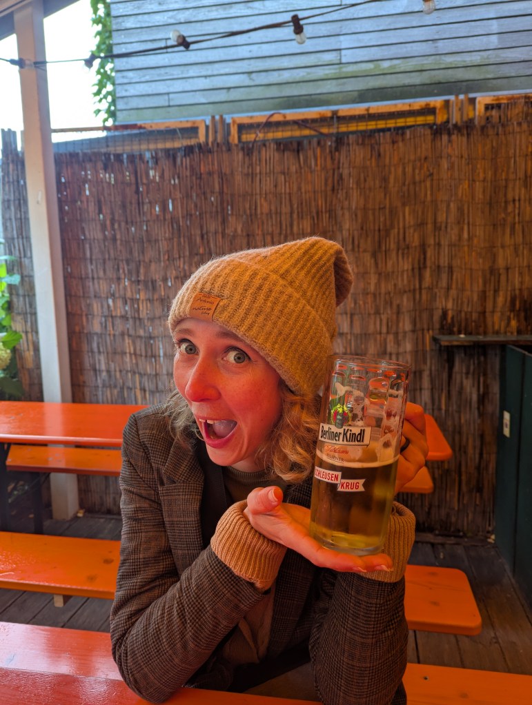 A woman wearing a beige beanie and a blazer is excitedly holding a glass of beer labeled 'Berliner Kindl' while sitting at an orange picnic table in a casual outdoor setting.