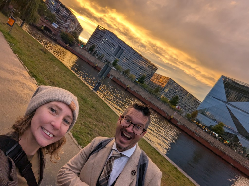 A man and a woman taking a selfie by the river at sunset, with modern buildings in the background and a clear sky filled with clouds.