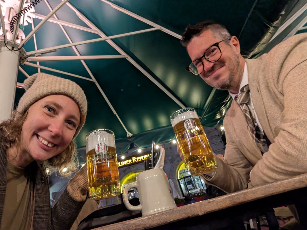 A man and woman sitting at a table in an outdoor setting, each holding a large beer mug, smiling at the camera, with a green canopy overhead.