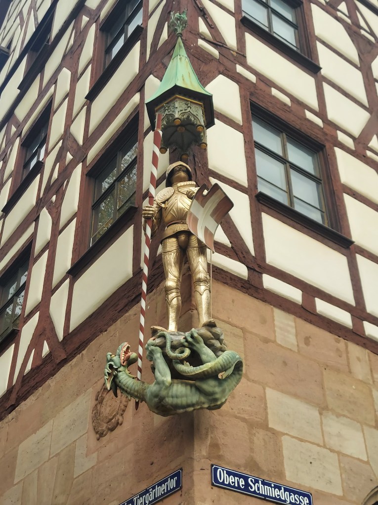 A decorative sign featuring a golden knight standing atop a green dragon, with a lantern above and a striped pole beside it, mounted on a corner of a timber-framed building.
