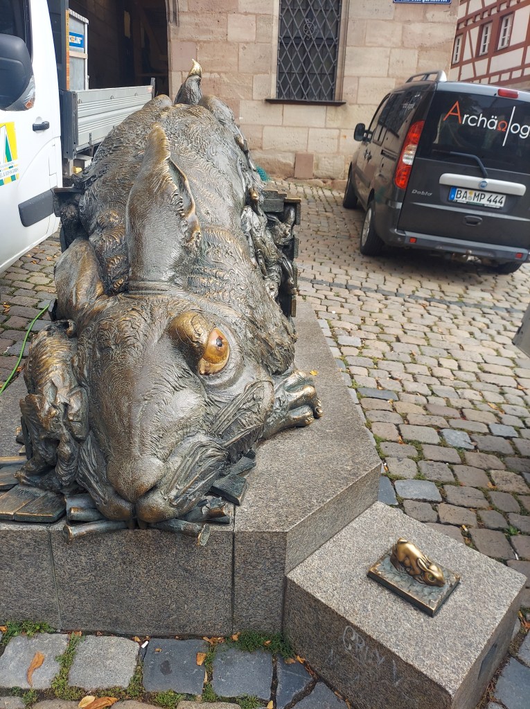 A large bronze sculpture of a rabbit, positioned on a stone pedestal, with intricate details showcasing its fur and features. There is a smaller bronze figure of a rabbit on a lower stone base nearby. The background includes cobblestone streets and parked vehicles.