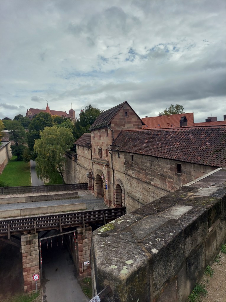 View of a historic stone structure with a bridge and pathway in the foreground, set against a cloudy sky and surrounded by greenery.