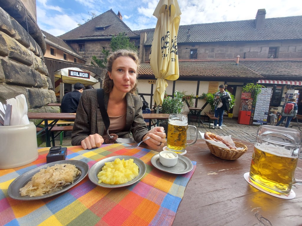 A woman seated at an outdoor table with traditional German dishes, including sauerkraut and mashed potatoes, alongside two glasses of beer. The setting features rustic architecture and other patrons in the background.
