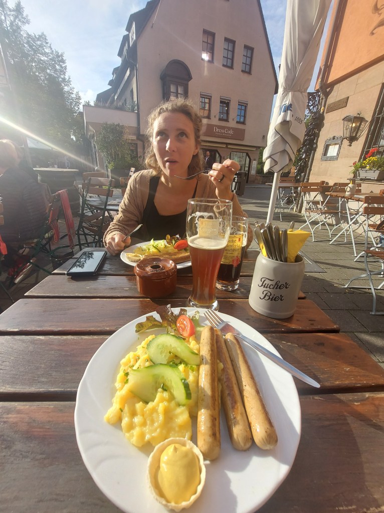A woman sitting at an outdoor table, looking up thoughtfully while holding a spoon. In front of her is a plate of food including creamy mashed potatoes, cucumber slices, sausages, and a small serving of mustard. A glass of beer is also visible on the table.