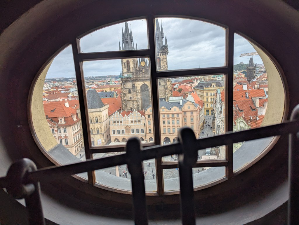 View of Prague's Old Town Square through an arched window, featuring the Church of Our Lady before Tyn and colorful rooftops under a cloudy sky.
