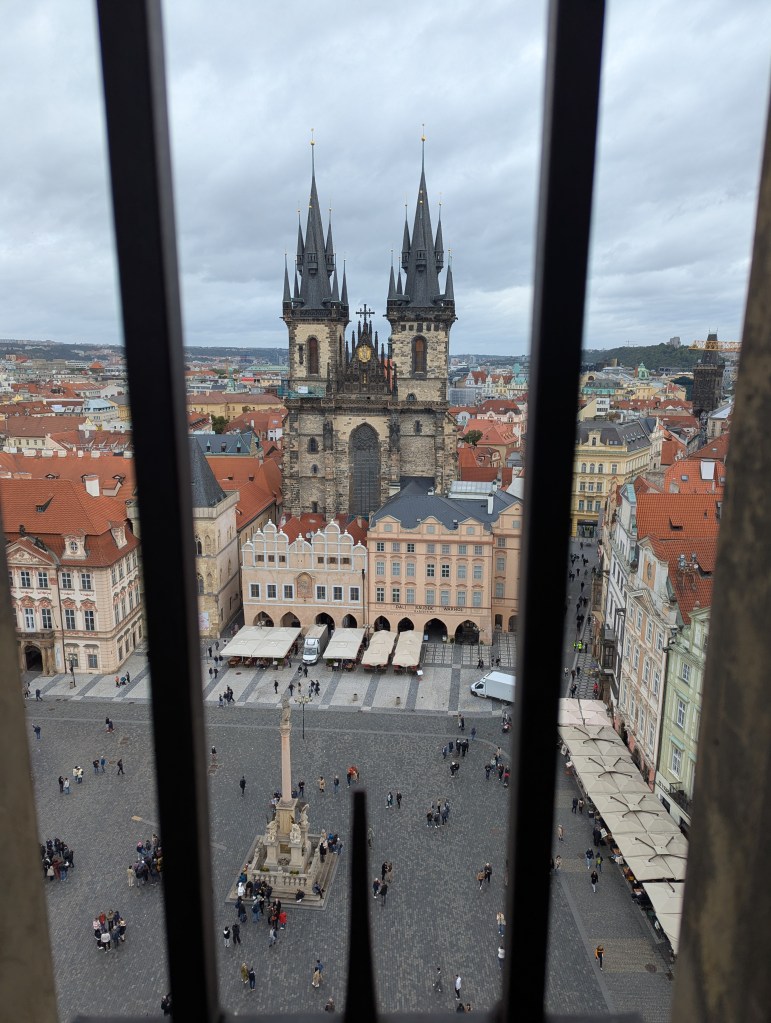 View of the Church of Our Lady before Týn with its distinctive spires, framed by a metal railing, overlooking a bustling square with cobblestone streets and people gathered below.