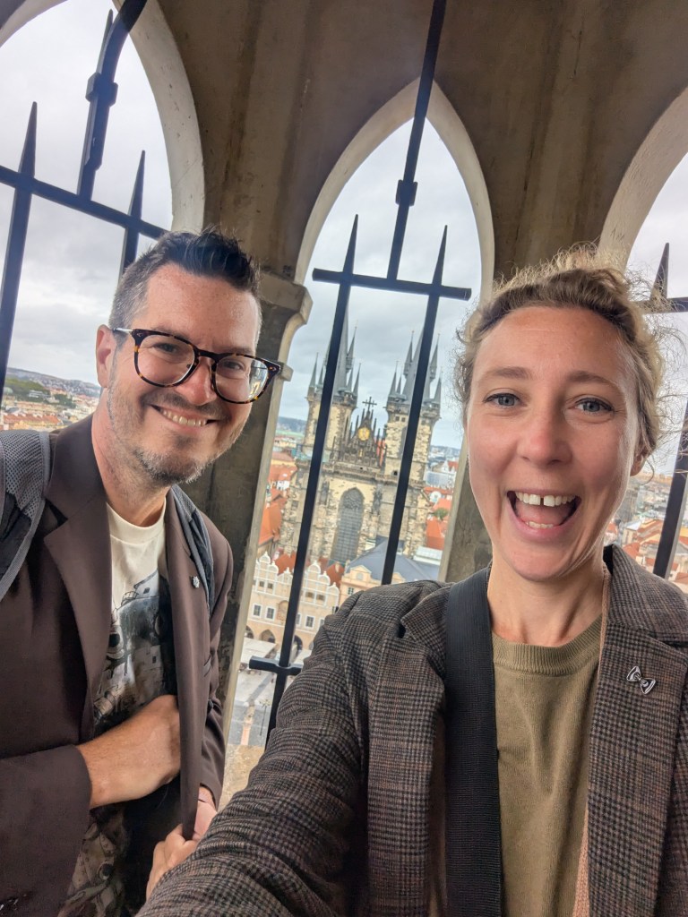 Two smiling individuals pose for a selfie in an archway, overlooking a cityscape with historic buildings in the background.