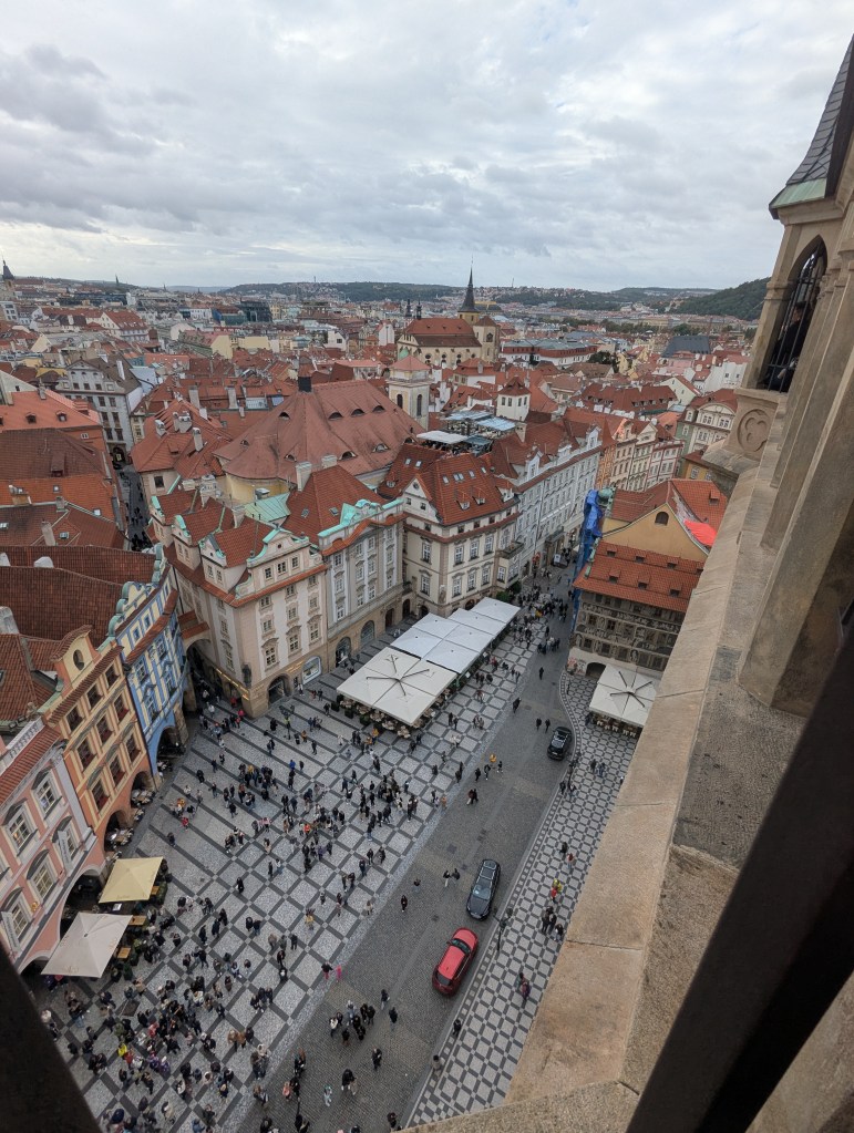 Aerial view of a bustling square in a historic city, featuring red-roofed buildings and a patterned cobblestone plaza filled with people. Numerous outdoor cafe tables and vehicles can be seen, under a cloudy sky.