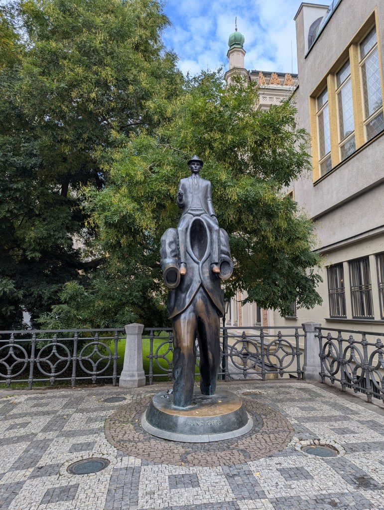 A bronze statue of a man in a suit, holding a book, on a pedestal, surrounded by trees and a decorative railing.