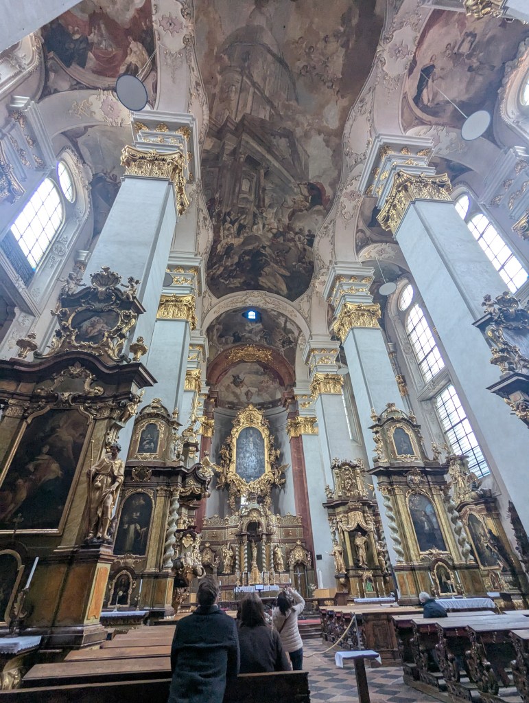 Interior view of a grand cathedral showcasing ornate architecture, high ceilings with frescoes, and detailed altars. Several visitors are seen observing the artistic elements.