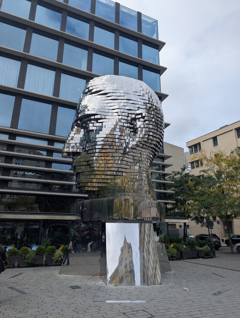 A large, reflective sculptural head made of metallic strips, placed in front of a modern glass building, surrounded by greenery and people.