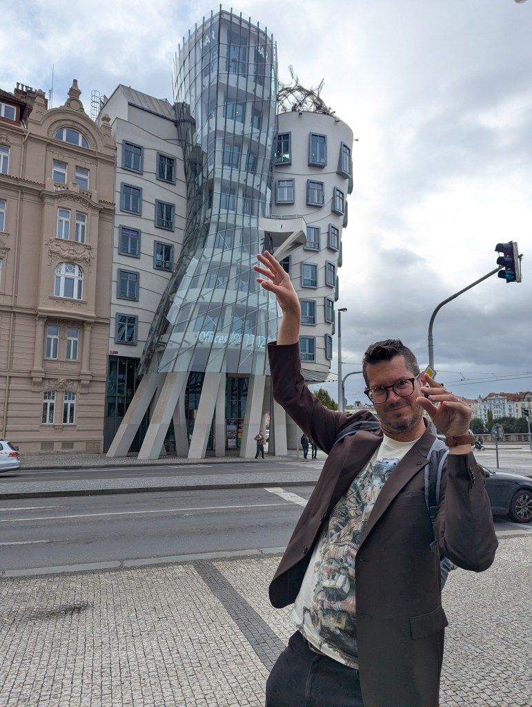 A man poses playfully in front of the unique Dancing House building in Prague, showcasing its curved glass architecture and contrasting traditional structures.