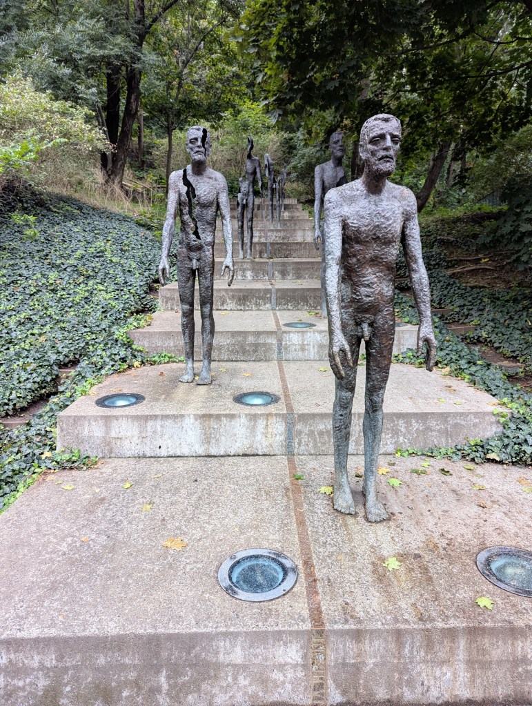A series of bronze sculptures resembling human figures ascending a set of concrete steps, surrounded by greenery.