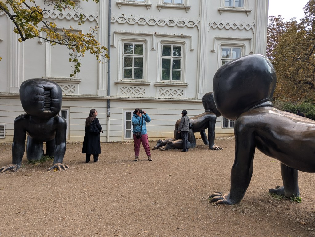 A group of large, stylized bronze sculptures resembling humanoid figures with oversized heads and exaggerated features, situated in a garden setting near a historical building. Two individuals are visible, one taking a photograph and the other standing beside a sculpture.