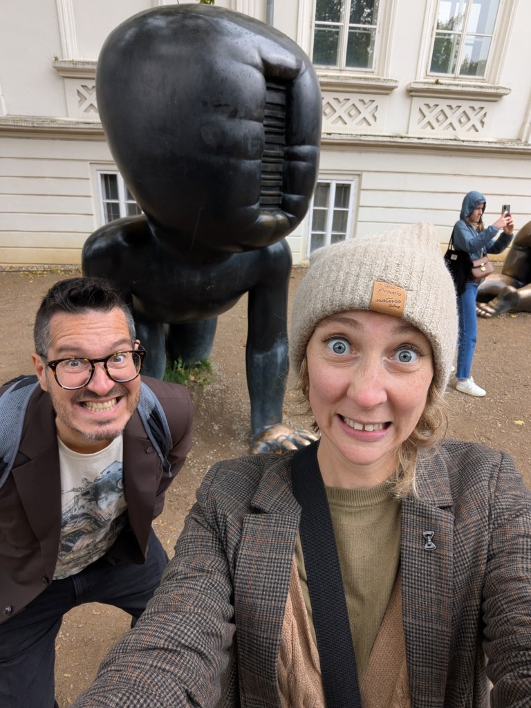 A selfie of two smiling individuals in front of a large, dark sculpture resembling a figure with a distorted face in a park setting.