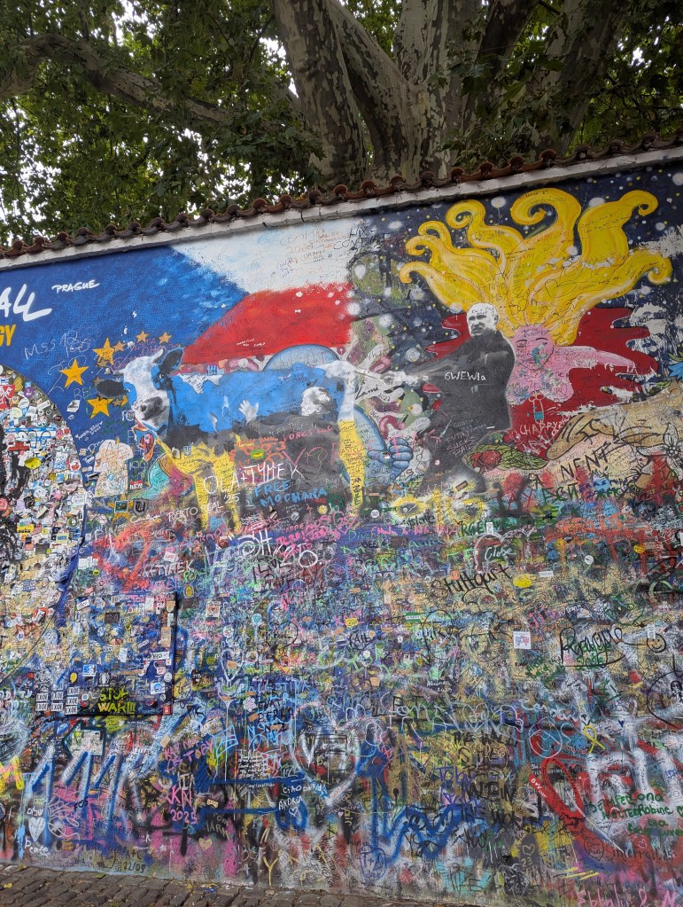 A colorful mural featuring vibrant graffiti art, including abstract shapes, text, and portraits, with a tree partially visible at the top.