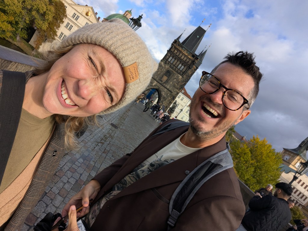 A joyful couple poses for a selfie on a cobblestone street with a historic tower in the background, smiling and wearing warm clothing.
