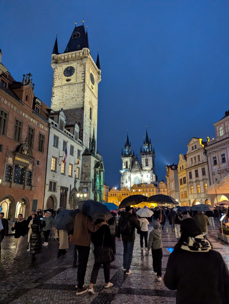 A bustling night scene in a historic square with a clock tower and gothic spires, where people are walking under umbrellas on a cobblestone street.